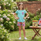 A young girl stands in a garden wearing a light blue t-shirt with a red and blue plaid chest pocket and matching plaid shorts. A small table next to her holds a 'Bean' book and a small Union Jack flag.