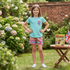 A young girl stands in a garden wearing a light blue t-shirt with a red and blue plaid chest pocket and matching plaid shorts. A small table next to her holds a 'Bean' book and a small Union Jack flag.