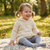 A young child with pigtails sits smiling on a patterned blanket in a sunlit park, wearing a cream knit hooded long-sleeve top and gray leggings.