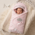 A sleeping newborn baby wrapped in a light pink plush swaddle blanket with lace trim, featuring a crown applique and the word 'PRINCESS' on the hood. The swaddle is displayed inside a white wicker basket on a cream cable-knit blanket.