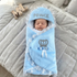 A sleeping newborn baby wrapped in a light blue plush swaddle blanket with lace trim, featuring a hood with 'PRINCE' embroidered on it, a silver crown motif, and a blue and white ribbon bow. The baby is lying on a gray quilted blanket.