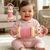 Baby in pink outfit holding a toy with a smiling expression, with another toy in the background.