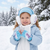 Young girl smiling outdoors in a snowy, pine tree background, wearing a light blue winter jacket with Disney Frozen Elsa earmuffs and matching fingerless gloves holding a snowflake ornament.