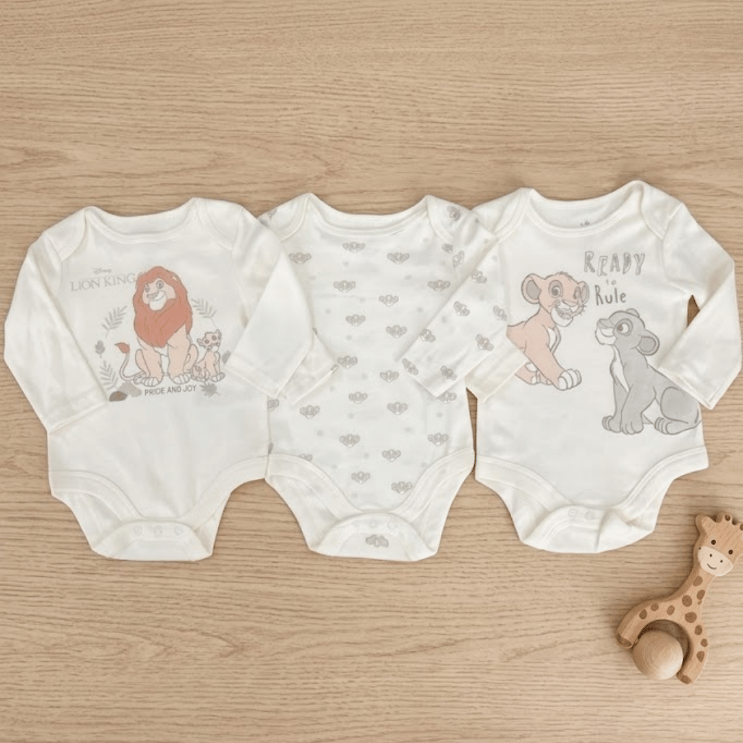 Three baby bodysuits with cartoon designs on a wooden surface, accompanied by a wooden toy.