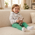 A young child sitting on a cream-colored boucle sofa, wearing a light gray hoodie, green ribbed leggings, and white socks while holding a small gray plush elephant toy.