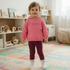 A toddler wearing a pink hooded sweatshirt with a small chest graphic and kangaroo pocket, paired with maroon ribbed leggings and white socks, standing in a brightly colored playroom with foam alphabet mats on the floor, a wooden toy kitchen, and a bookcase filled with children's books.