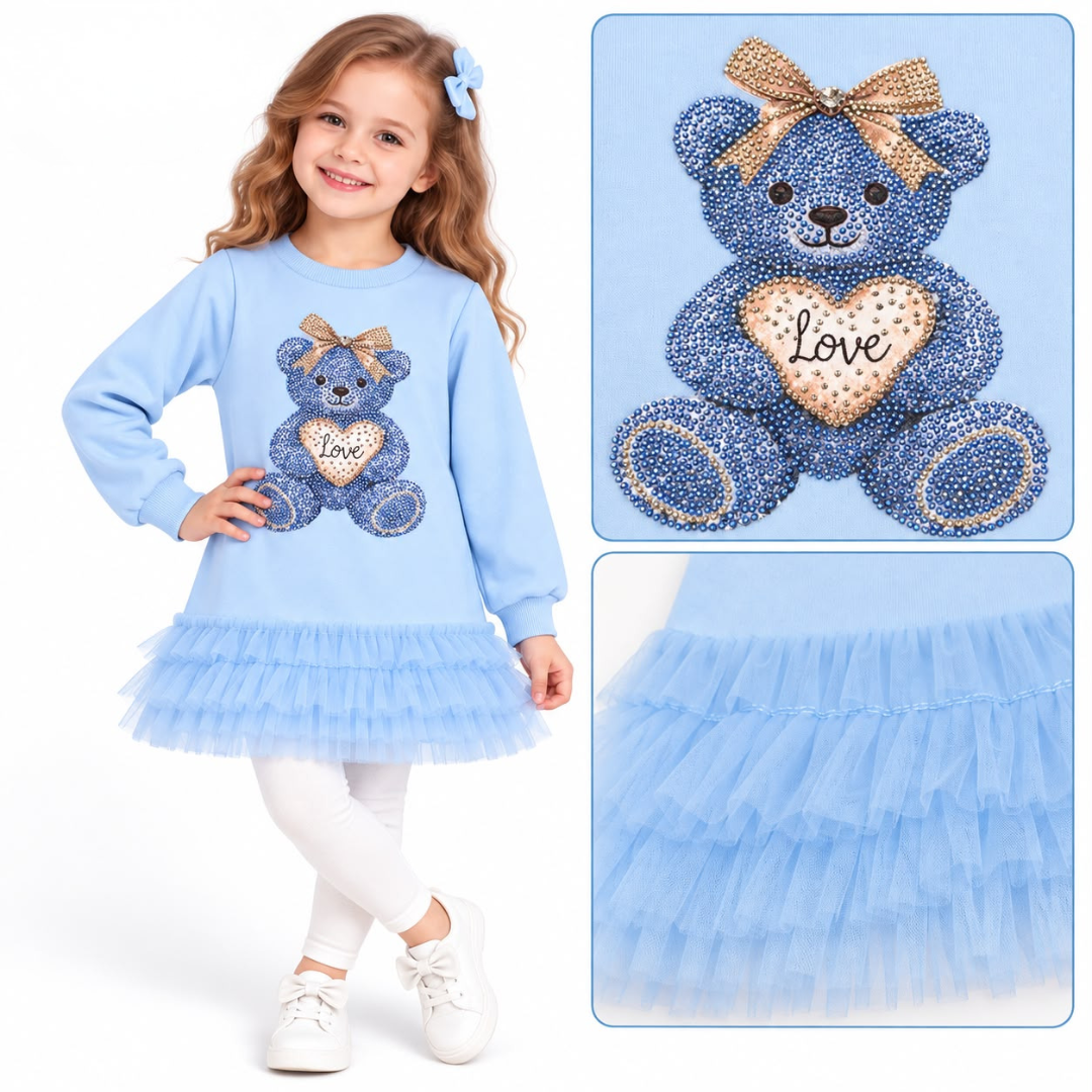 A young girl modeling a light blue long-sleeve tulle dress with a rhinestone teddy bear graphic on the front. The dress features a layered tulle skirt and a matching teddy bear design with a gold bow and heart on the chest.