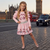 A young girl with blonde braided hair stands on a cobblestone street in London, wearing a pink long-sleeve dress with cherry sequin details. The background shows Big Ben and a red double-decker bus.