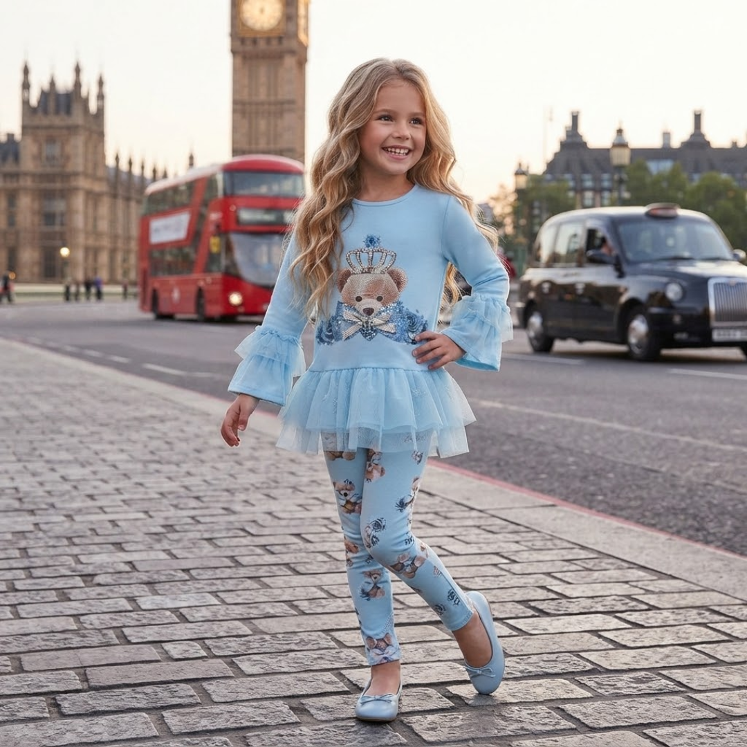 A young girl smiling while walking on a cobblestone street in London, wearing a light blue long-sleeve tunic top with a teddy bear graphic and matching light blue floral leggings.