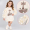 A young girl models a cream-colored long-sleeve dress featuring an embroidered Eiffel Tower and roses on the front, with sheer tulle sleeves decorated with cream satin bows. Two circular inset close-ups on the right show detailed views of the embroidered tower motif and a close-up of the bow on the sleeve.
