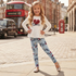 A young girl stands on a city street wearing a white long-sleeve top with a red heart and 'Love' graphic, paired with light blue leggings featuring a dark script and floral print. A red double-decker bus and London street elements are visible in the background.