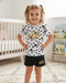 A young girl with curly blonde hair in pigtails stands in a nursery wearing a white short-sleeve t-shirt and black shorts. The t-shirt features black polka dots, cartoon character prints, and gold 'Disney 101 Animal Kingdom' text.