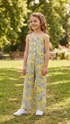 A young girl stands outdoors on a grassy lawn wearing a yellow sleeveless jumpsuit with a gray palm leaf pattern and white sneakers.