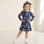 A young girl stands with hands on hips wearing a navy blue long-sleeve dress with a colorful cartoon character print, a matching navy bow headband, and white sneakers in a bright, minimalist indoor setting.