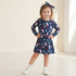 A young girl stands with hands on hips wearing a navy blue long-sleeve dress with a colorful cartoon character print, a matching navy bow headband, and white sneakers in a bright, minimalist indoor setting.