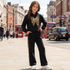 A young girl stands on a London street wearing a black two-piece tracksuit with a large gold butterfly print on the hoodie and gold leopard-print side stripes on the pants, paired with white sneakers.
