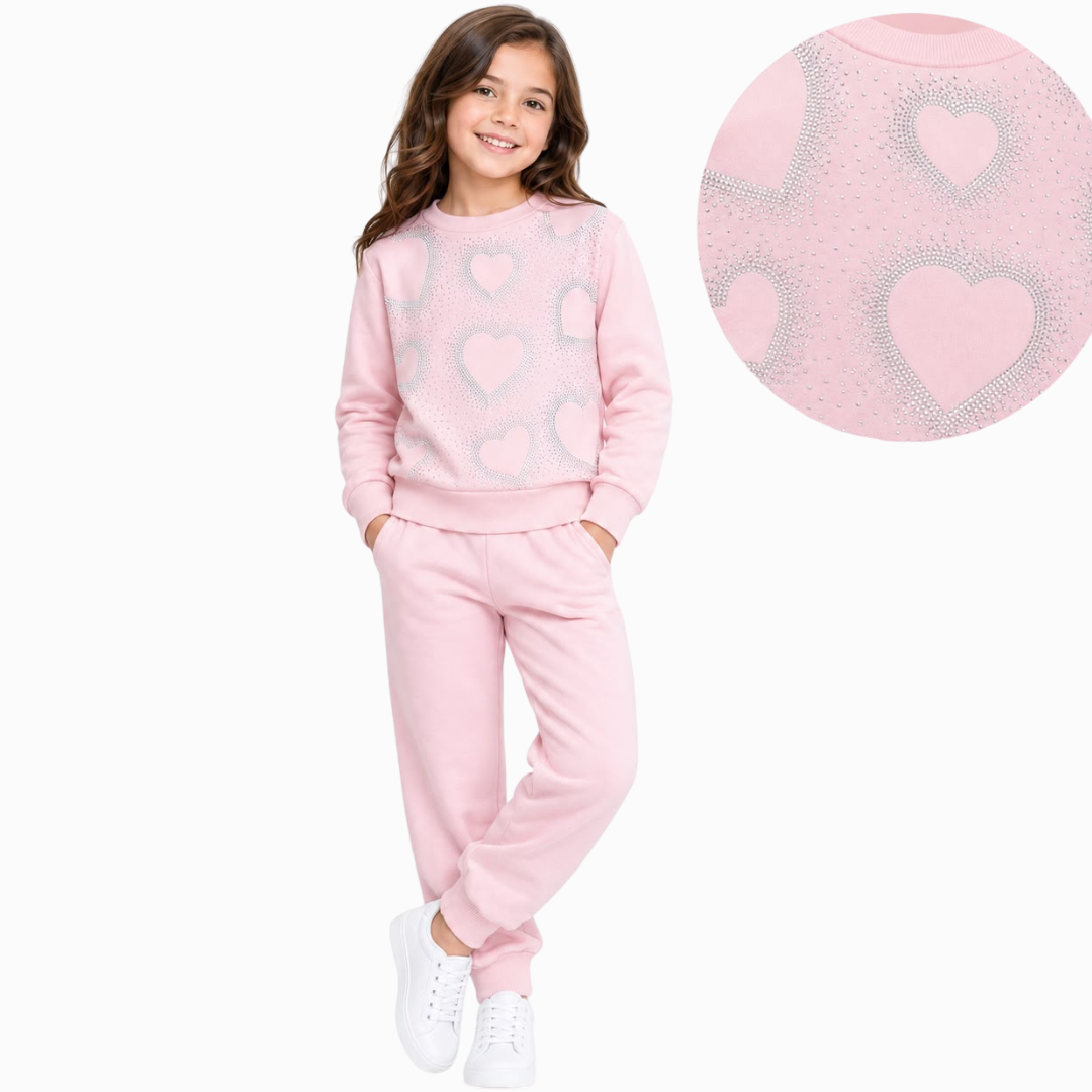 A young girl modeling a light pink two-piece sweatpants set with silver glittery heart patterns, shown against a plain white background with a circular detail inset of the heart pattern.