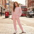 A young girl stands on a city street wearing a light pink sweatshirt and matching jogger pants with a subtle tonal heart pattern. The background shows a red double-decker bus, a black cab, and a red post box.