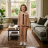 A young girl stands in a living room wearing a matching beige ribbed knit two-piece outfit with black trim and white sneakers. The set includes a long-sleeve top with a black button placket and matching pants with black side stripes.