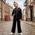 A young girl stands on a cobblestone street wearing a black zip-up hoodie with rhinestone embellishments on the chest and matching black wide-leg trousers, paired with white sneakers.