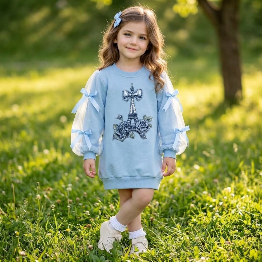 A young girl stands in a grassy park wearing a light blue sweatshirt with sheer balloon sleeves, blue bow details, and a Paris Eiffel Tower graphic on the front.