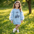 A young girl stands in a grassy park wearing a light blue sweatshirt with sheer balloon sleeves, blue bow details, and a Paris Eiffel Tower graphic on the front.