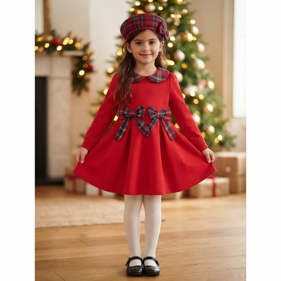 A young girl wearing a festive red long-sleeve dress with a plaid collar and waist bow, matching plaid beret, white tights, and black Mary Jane shoes, standing in a holiday-themed room with a decorated Christmas tree in the background.