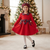 A young girl wearing a festive red holiday dress with a red and blue plaid collar, matching plaid beret, and a plaid bow at the waist, standing in a decorated living room with a Christmas tree in the background.