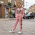 A young girl stands on a cobblestone street wearing a pink matching sweatshirt and jogger set with black leopard-print butterfly and bow embellishments. The background shows a brick street with a black cab and buildings.