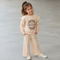 A young girl with a brown ponytail stands outdoors on a gray concrete surface, wearing a light peach-beige two-piece lounge set consisting of a sweatshirt with ruffled sleeves and a matching pair of wide-leg pants. The sweatshirt features a circular graphic with the word 'DREAMER' in the center.