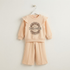 A two-piece children's outfit hanging on a white hanger against a plain light beige wall. The set includes a long-sleeve sweatshirt with a large circular 'DREAMER' graphic on the front and matching wide-leg pants.