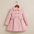A light pink children's double-breasted coat with a Peter Pan collar, decorative gold buttons, and matching bow details at the waist, hanging on a wooden hanger against a plain beige wall.