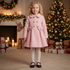 A young girl with curly blonde hair stands in a cozy holiday living room wearing a light pink knee-length coat with a double-breasted button closure and two large bow details at the waist, paired with white tights and dark red Mary Jane shoes. A decorated Christmas tree, fireplace with candles, and wrapped gifts are visible in the background.