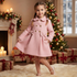 A young girl with braided hair stands in a cozy holiday room wearing a light pink knee-length coat with gold buttons, bow details, and a collar. A decorated Christmas tree, fireplace with candles, wrapped gifts, and a window are visible in the background.