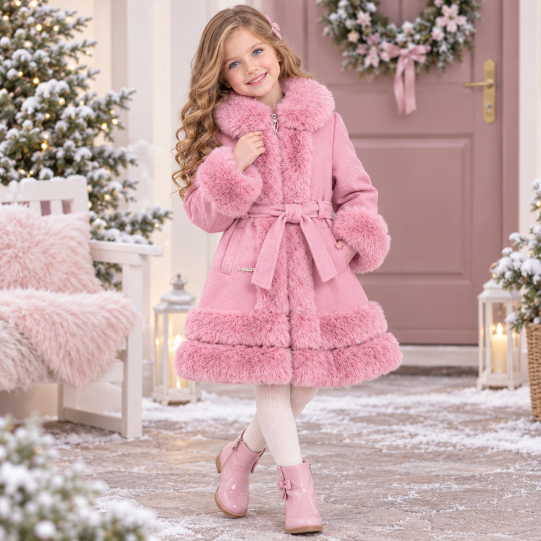 A young girl posing in a festive outdoor setting, wearing a pink winter coat with matching faux-fur trim, white tights, and pink boots.