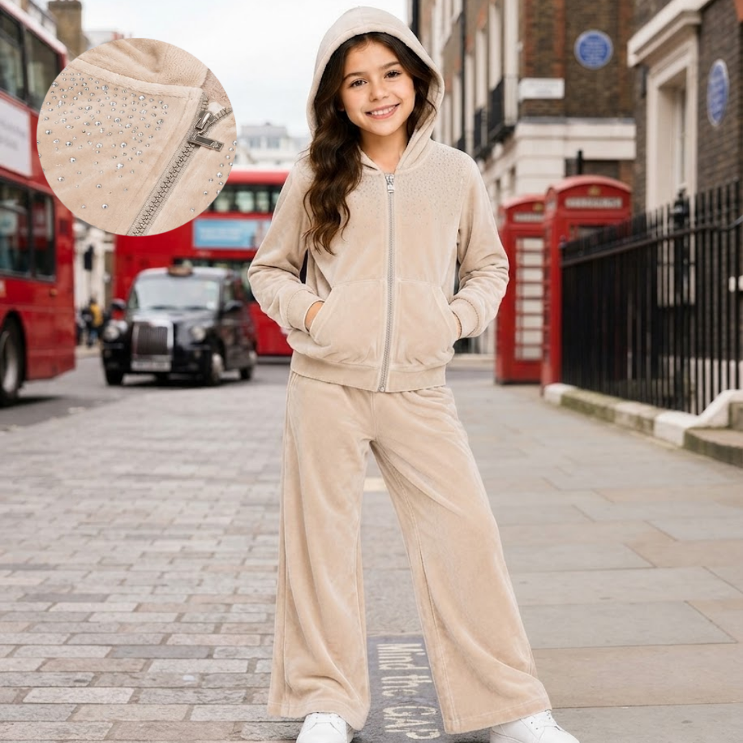 A young girl stands on a city street wearing a beige hooded zip-up jacket and matching wide-leg pants. The image includes a circular inset close-up showing a matching beige embellished pouch with a silver zipper.