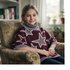 A smiling young girl sitting comfortably in an armchair indoors, wearing a cozy maroon knitted turtleneck poncho featuring a white star pattern and striped ribbed trim