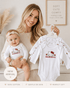 Woman holding a baby and a hanger with Hello Kitty-themed baby clothes in a cozy room.