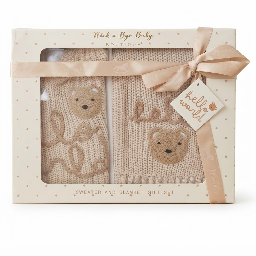 A boxed baby gift set from Rick a Bye Baby Boutique featuring a beige knit sweater and matching blanket with embroidered teddy bear faces and cursive text, presented in a cream gift box with a ribbon and tag.