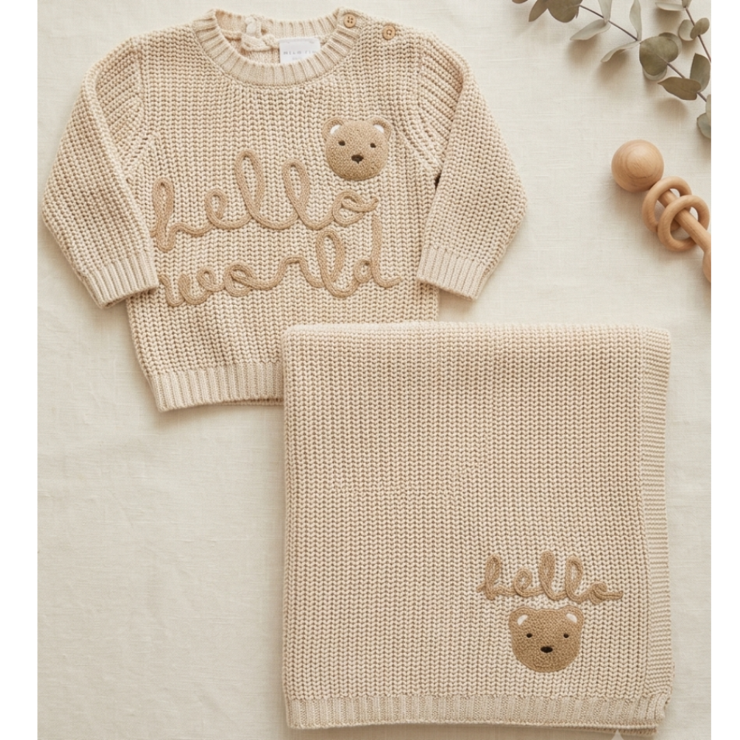 A beige knit baby sweater and matching blanket set, styled on a neutral fabric background with a wooden rattle and dried leaves. The sweater features 'hello world' written in cursive with a teddy bear face, and the matching blanket has a similar design.