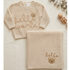 A beige knit baby sweater and matching blanket set, styled on a neutral fabric background with a wooden rattle and dried leaves. The sweater features 'hello world' written in cursive with a teddy bear face, and the matching blanket has a similar design.