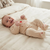 A baby lying on a cream-colored bed, wearing a matching beige footed sleeper with 'Mummy & Daddy' and a small red heart printed on the chest.