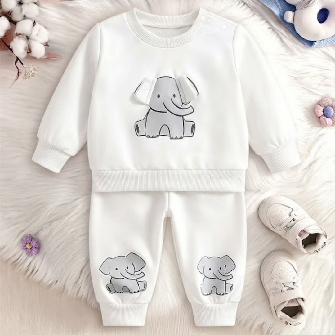A baby two-piece clothing set consisting of a white long-sleeve sweatshirt and matching pants, both featuring a gray cartoon elephant graphic. The set is displayed in a flat-lay arrangement on a white faux fur background, styled with baby shoes, cotton bolls, and small flowers.