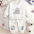 A baby two-piece clothing set consisting of a white long-sleeve sweatshirt and matching pants, both featuring a gray cartoon elephant graphic. The set is displayed in a flat-lay arrangement on a white faux fur background, styled with baby shoes, cotton bolls, and small flowers.