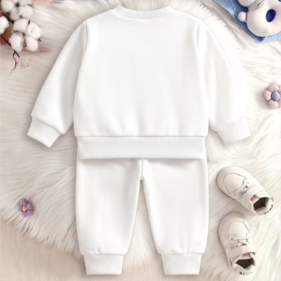 A baby two-piece sweatshirt and jogger pants set in solid white, laid flat on a white faux fur background with decorative props including cotton bolls, a plush toy, and baby shoes.
