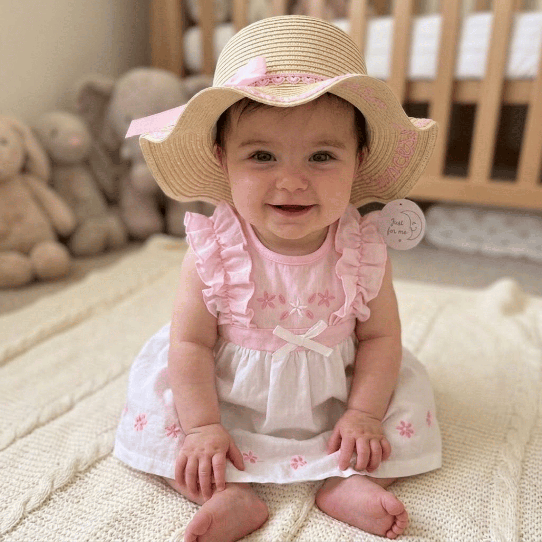 Baby in a pink dress and sun hat sitting on a crib with stuffed animals in the background