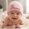 A smiling baby lying on a cream blanket wearing a light pink ribbed knit beanie with 'Princess' embroidered in dark pink script and a small crown detail.