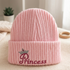 A pink ribbed knit beanie hat with a folded cuff, featuring an embroidered crown and the word 'Princess' in pink script on the front. The hat is displayed on a soft beige fabric surface with a small potted plant and wooden ring in the background.