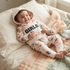 A baby lying on a bed wearing a floral print hooded sweatshirt and matching pants with the word 'GIRLS' printed across the chest. The scene includes a soft quilted blanket and a teddy bear.