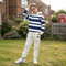 A young boy wearing a navy and white striped hoodie with a small black horse logo on the chest, paired with off-white sweatpants and white sneakers, standing on a grassy lawn in front of a brick house.